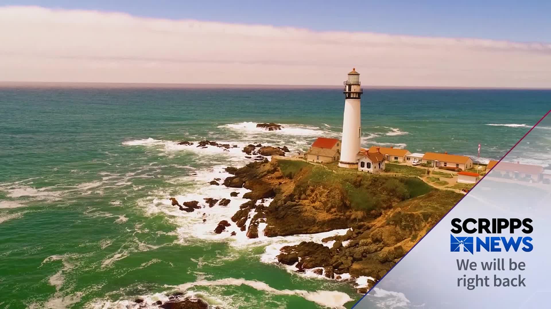 Waves crash against the rocky shoreline, while a white lighthouse stands tall on the cliff. The Scripps News graphic in the corner indicates a break in programming.
