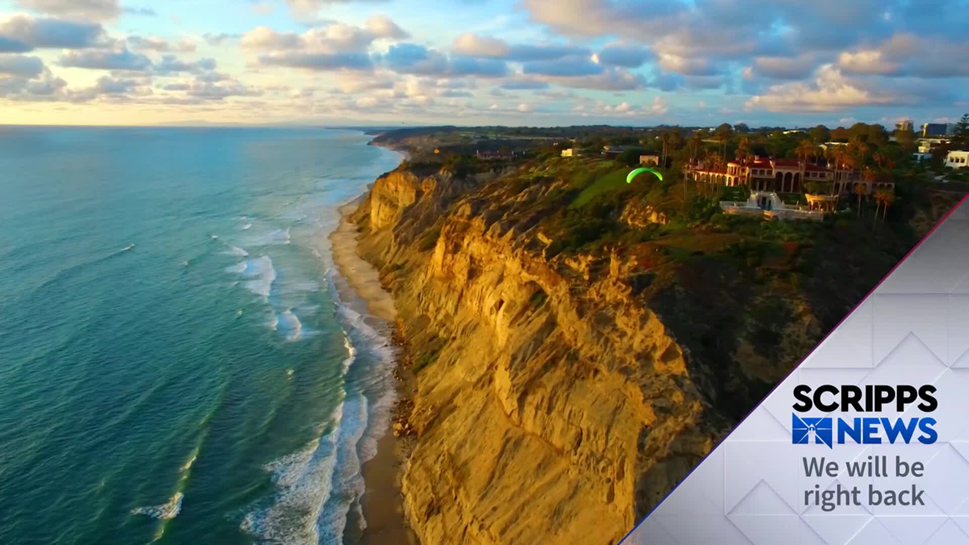 The turquoise ocean crashes against a sandy beach, and a paraglider floats above a cliffside mansion. The Scripps News graphic in the corner signals a break in the broadcast.
