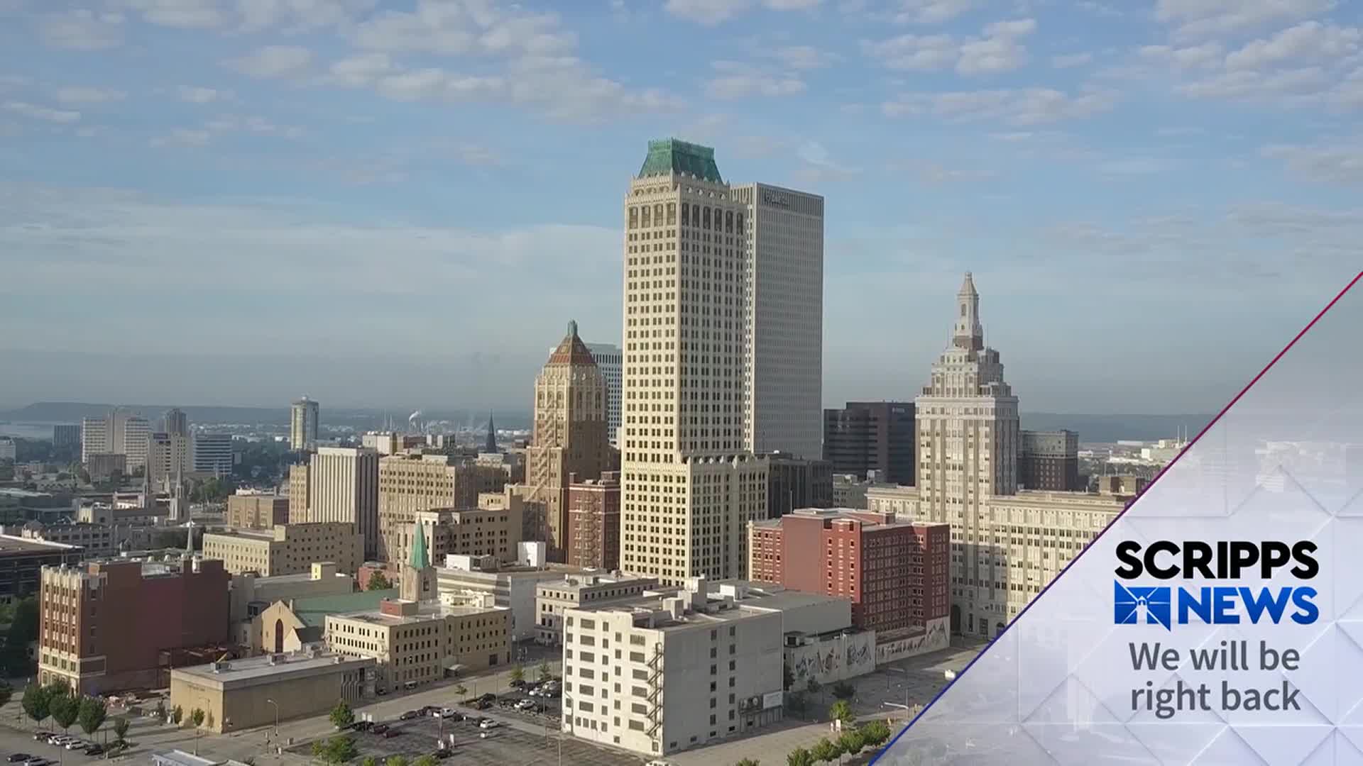 The camera looks down on a city skyline, with tall buildings dominating the view under a hazy sky. A Scripps News graphic is in the lower right corner, indicating a break in programming.
