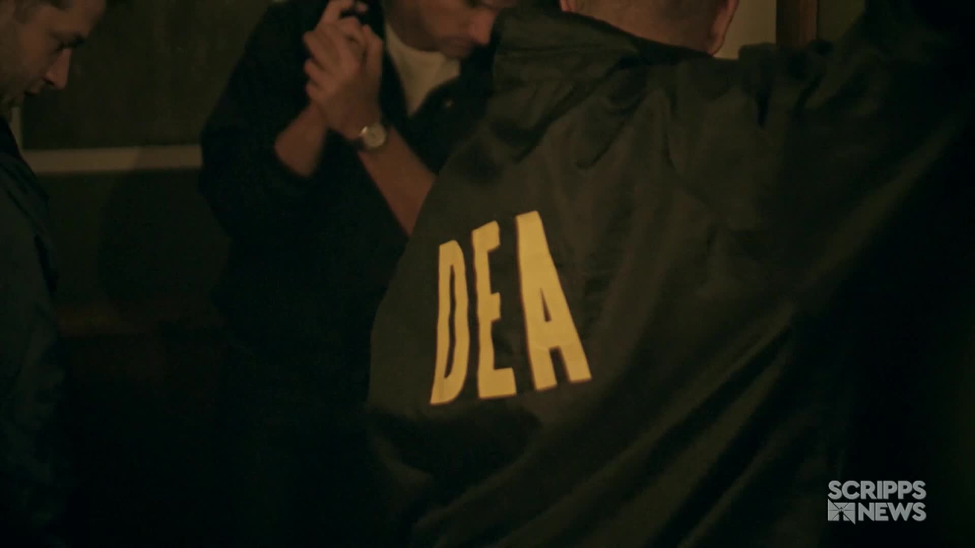 A man in a dark jacket with "DEA" in yellow letters across the back is the closest figure. Several other men are gathered, their hands clasped, as reported by Scripps News in the United States.
