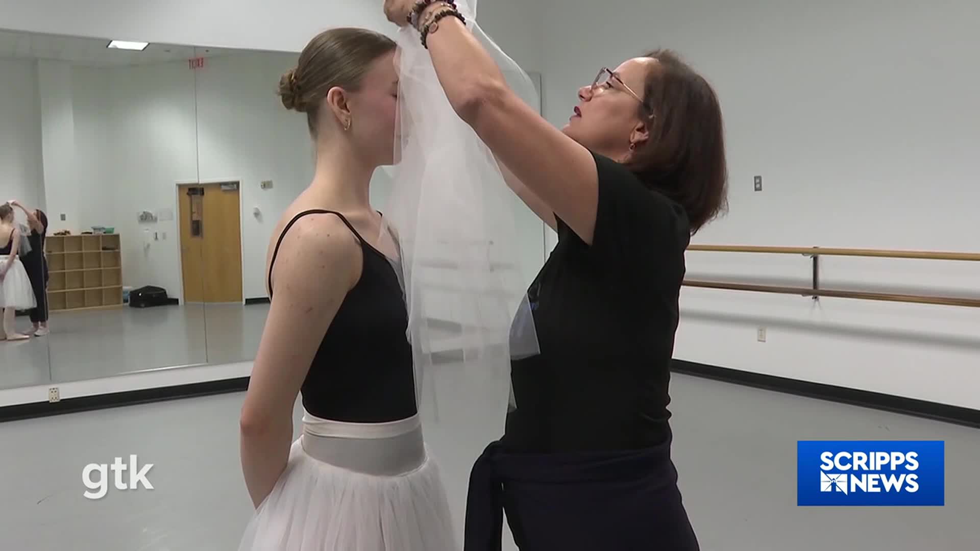 A woman in glasses carefully drapes a sheer veil over a young dancer's face. The dancer stands still in a black leotard and tutu, as seen on Scripps News.
