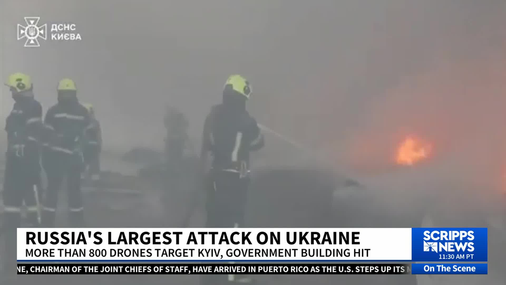 Firefighters in dark uniforms and yellow helmets are spraying water toward a bright orange blaze, obscured by thick smoke. The scene is being reported by Scripps News as Russia's largest attack on Ukraine.
