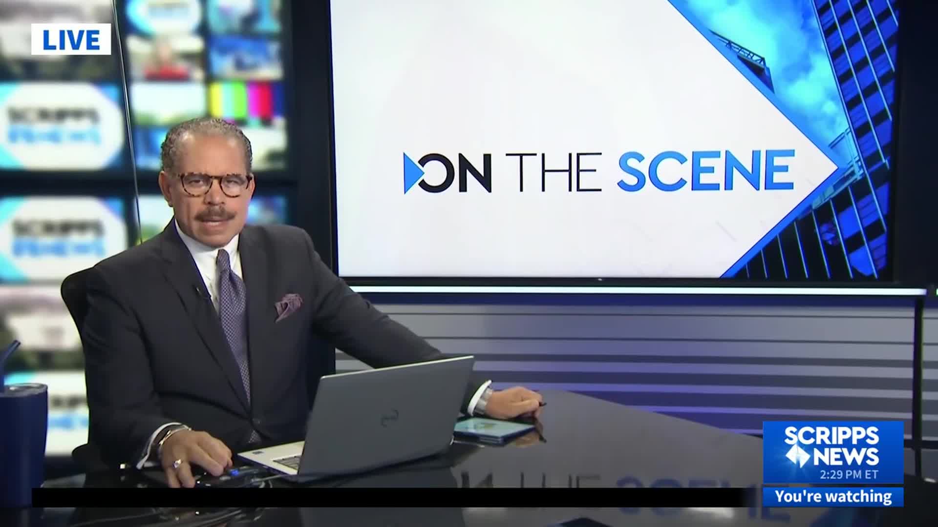 A man in a dark suit sits at a desk, looking directly at the camera. Behind him, the Scripps News logo and "ON THE SCENE" are displayed on a large screen.

