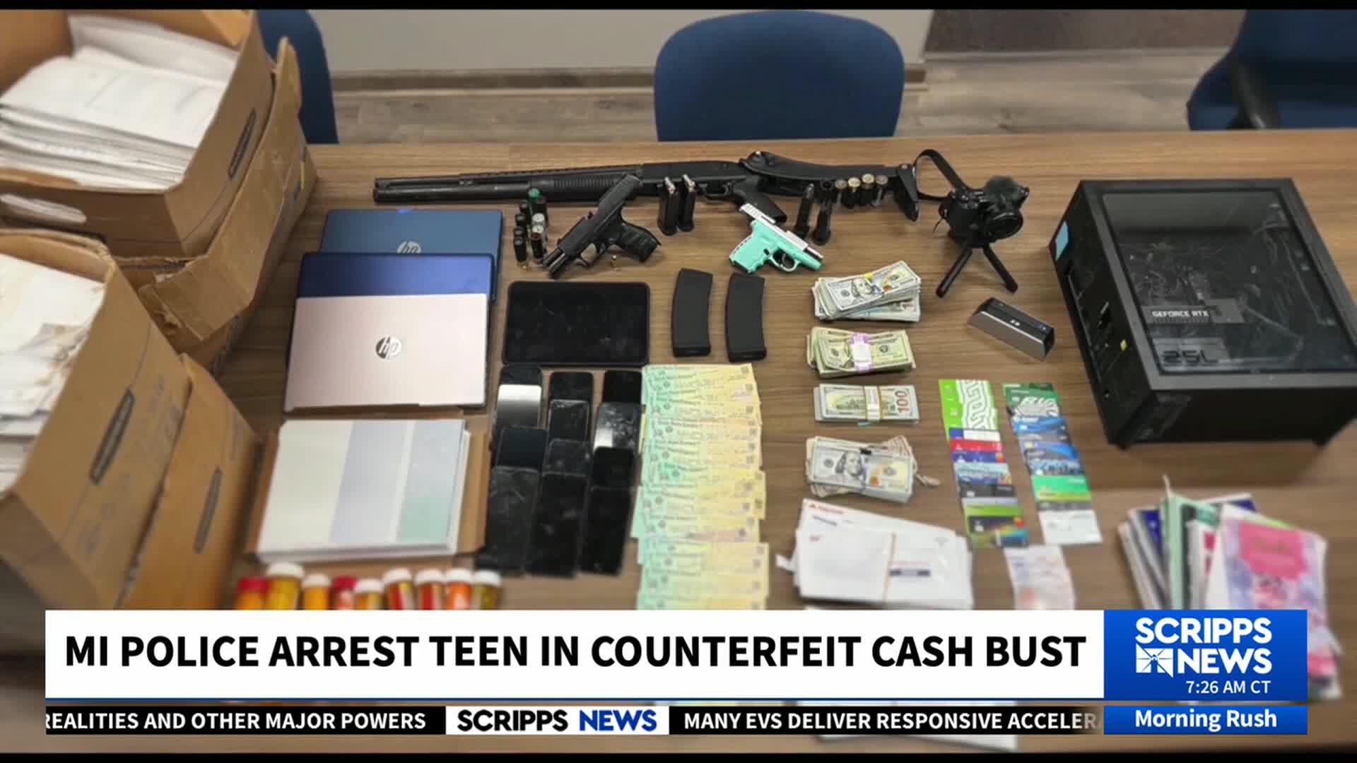 A table displays seized items, including a long gun, laptops, and stacks of cash, as reported by Scripps News. Numerous smartphones and a computer tower are also laid out, suggesting a recent police bust in Michigan.
