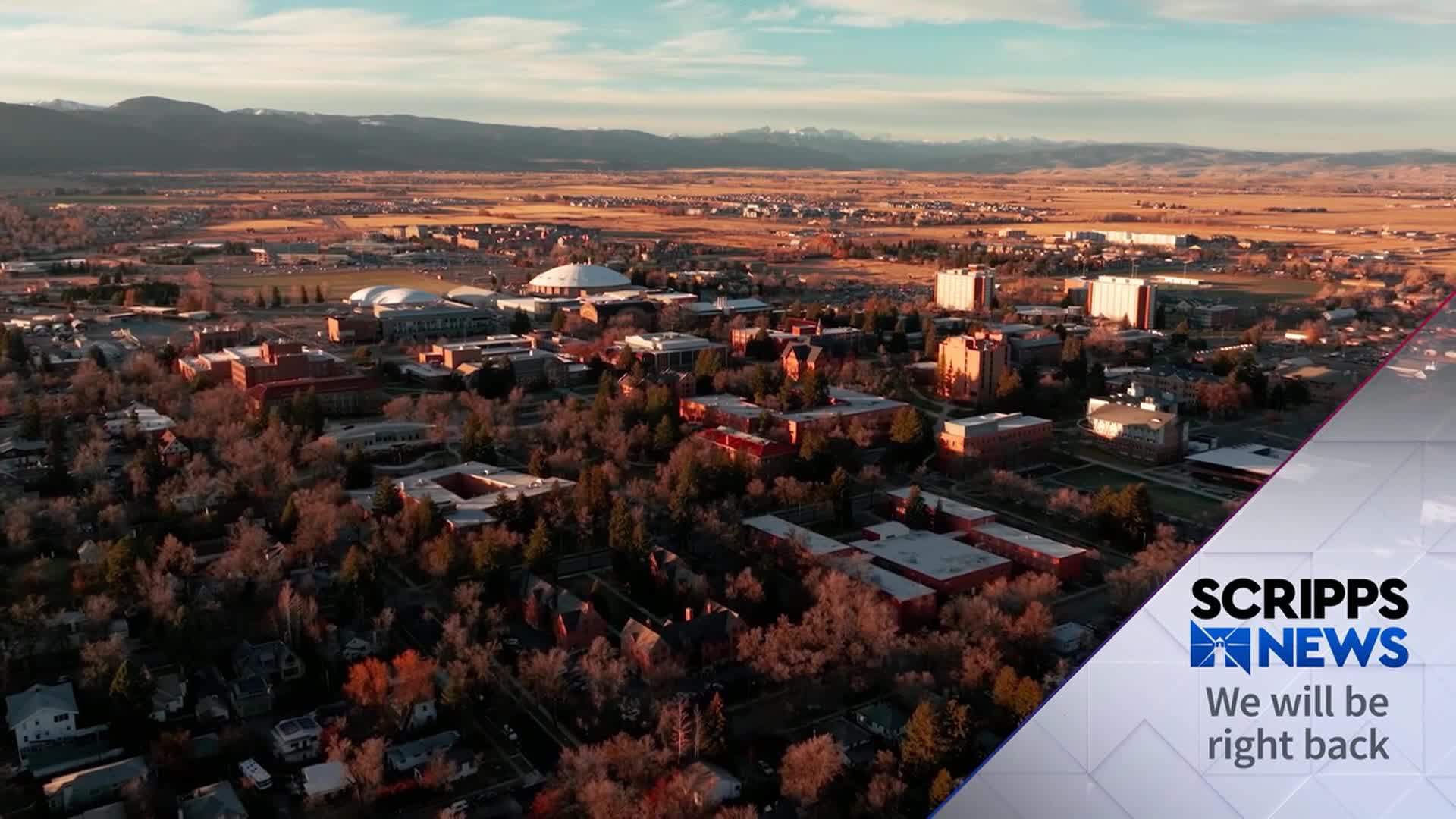 The sun is hitting the tops of buildings in a sprawling campus, casting long shadows across the grounds. Beyond the campus, the landscape stretches toward a mountain range under a clear sky, as Scripps News prepares to return.
