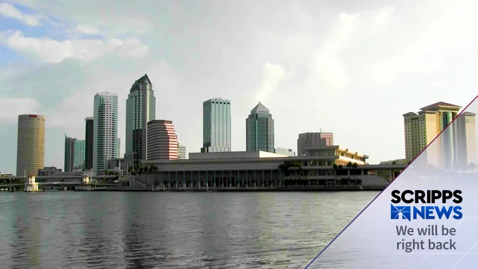 The Tampa, Florida skyline rises above the water, with a Scripps News graphic in the foreground. The sky is overcast, but the buildings still catch the light. 
