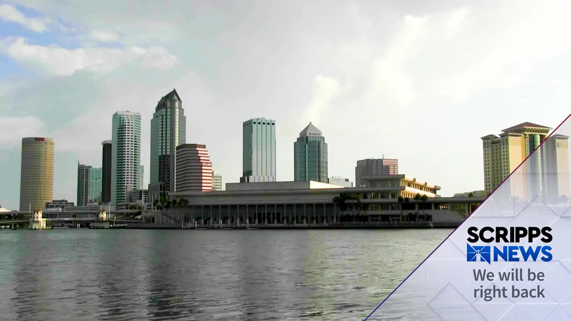 The Tampa skyline rises from the water, a mix of tall buildings against a cloudy sky. A Scripps News graphic in the corner signals a break in the broadcast.

