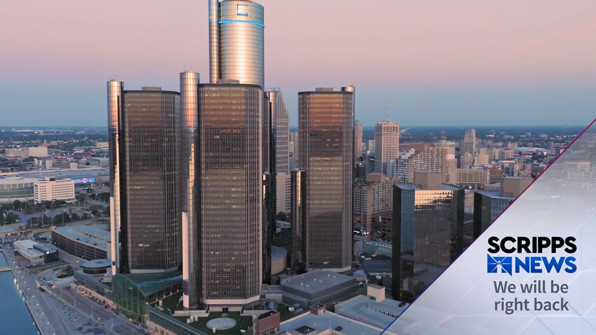The Detroit skyline, bathed in the soft light of dusk, is dominated by the gleaming towers of the Renaissance Center. A Scripps News graphic in the foreground indicates a break in programming.
