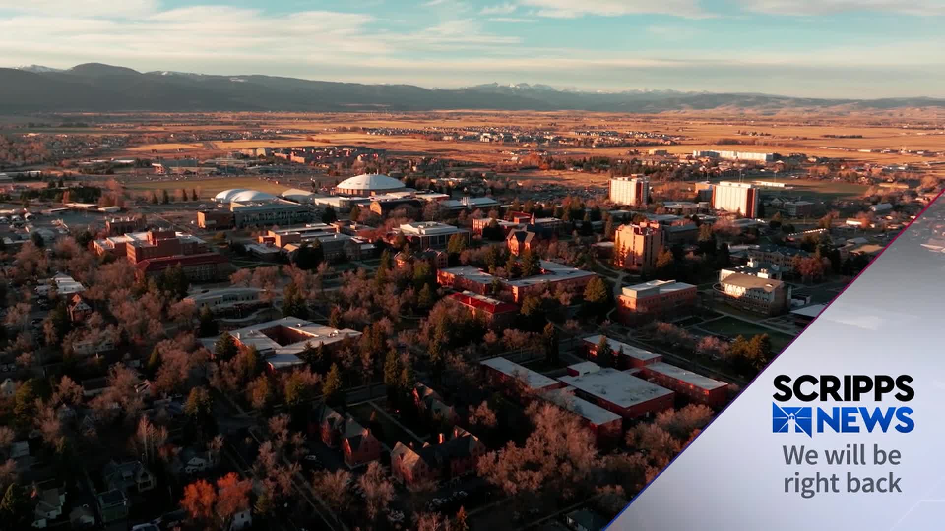 Sunlight bathes a university campus, its red-brick buildings and dome-shaped structures standing out against the distant mountains. A Scripps News graphic appears, indicating a break in programming.
