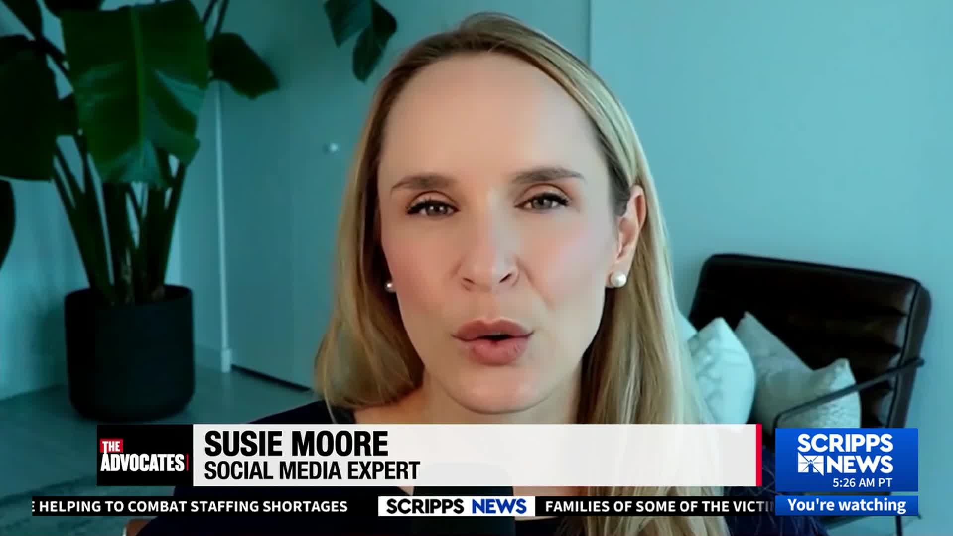 Susie Moore, a social media expert, is speaking on Scripps News, her mouth moving as she addresses the camera. Behind her, a large potted plant and a black chair are visible.
