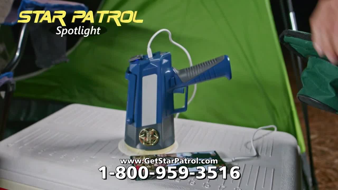 A blue and gray spotlight, shaped like a futuristic gun, sits on a cooler next to a phone. A person's hand is reaching for a green camping chair in the background.
