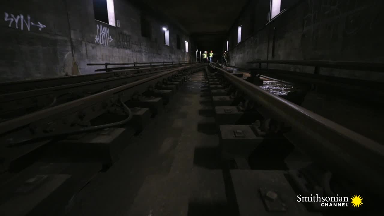 The tracks stretch into the darkness of a subway tunnel, where a few figures in safety vests are working in the distance. Water glistens on the concrete between the rails, reflecting the dim overhead lights.

