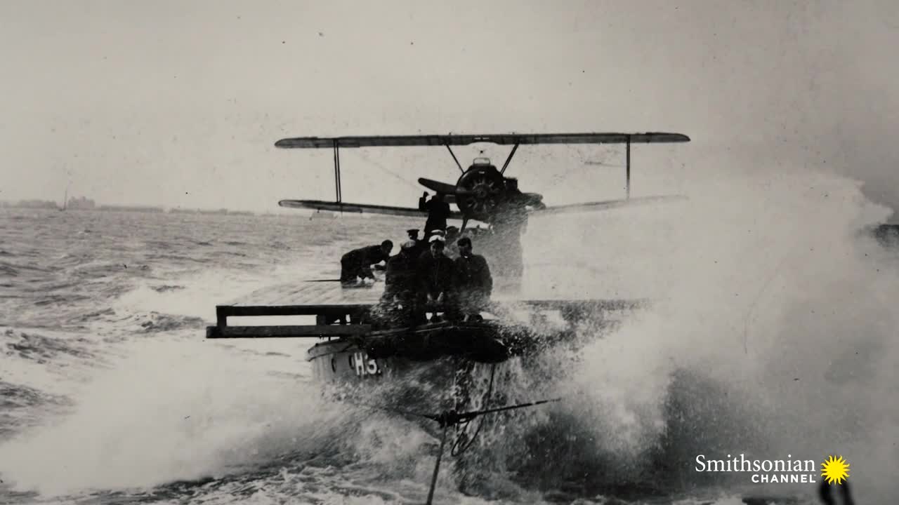 A biplane sits atop a boat, its wings silhouetted against the gray sky. The bow of the boat cuts through the choppy water, sending a wall of spray over the front.
