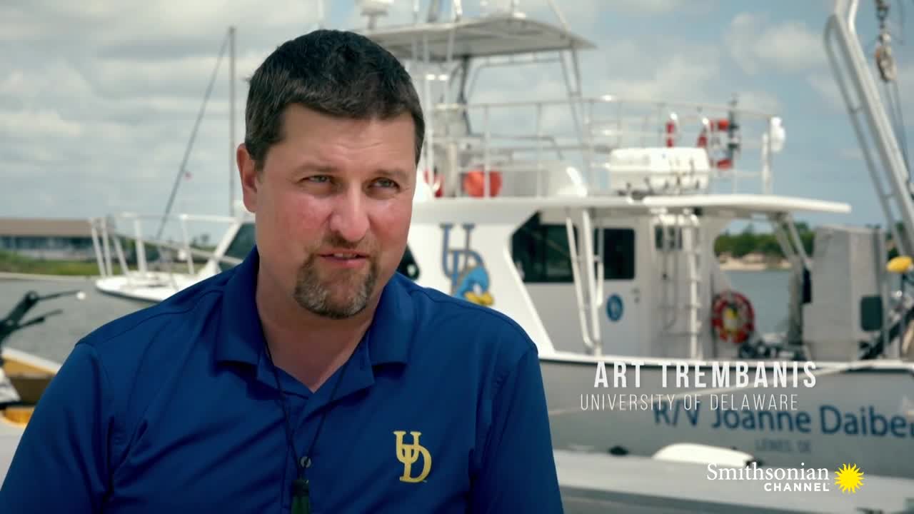 A man wearing a blue shirt with a University of Delaware logo is speaking. Behind him, a boat named "R/V Joanne Daiber" is docked, and the Smithsonian Channel logo is in the corner.
