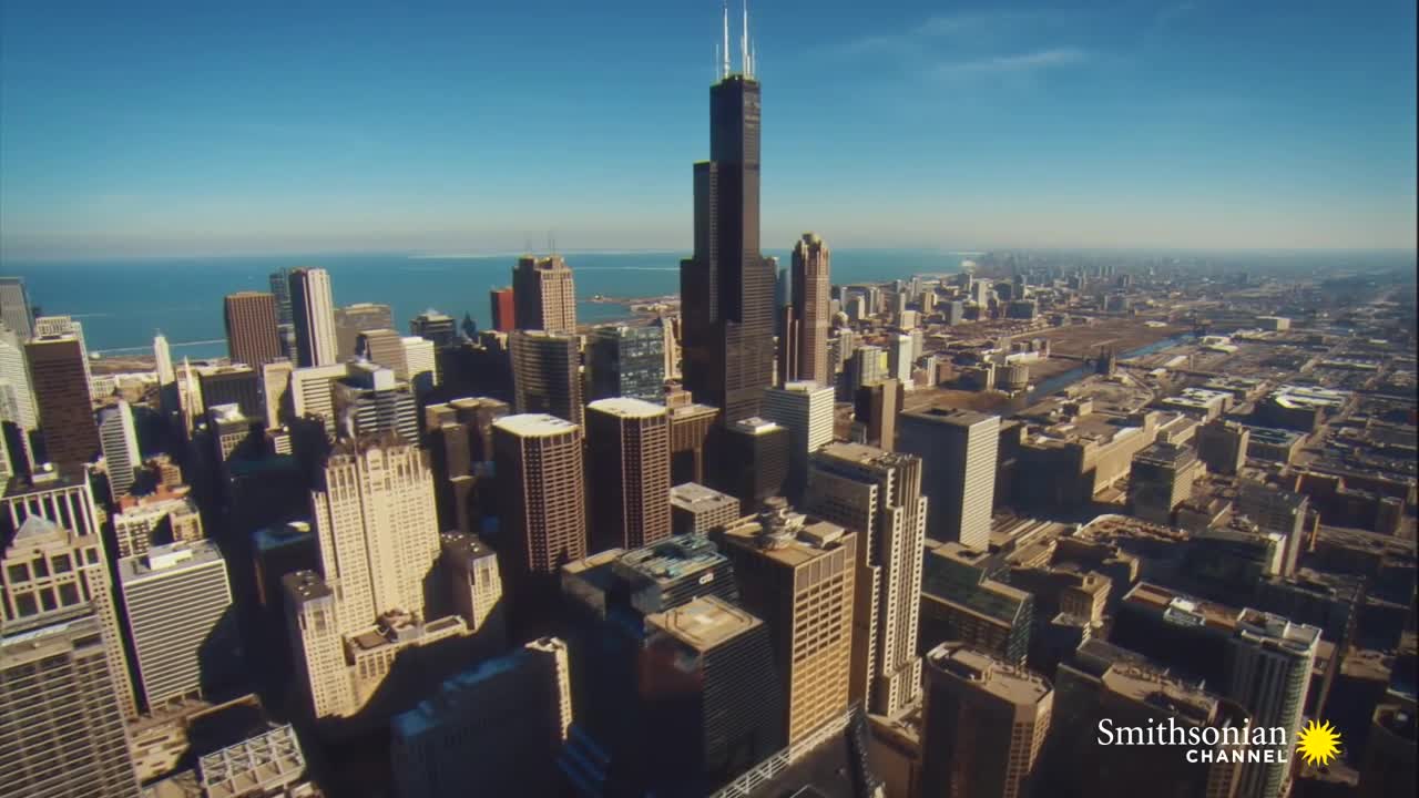 The towering Willis Tower dominates the Chicago skyline, its dark facade contrasting with the lighter buildings surrounding it. Beyond the city, the vast expanse of Lake Michigan shimmers under a clear, blue sky.
