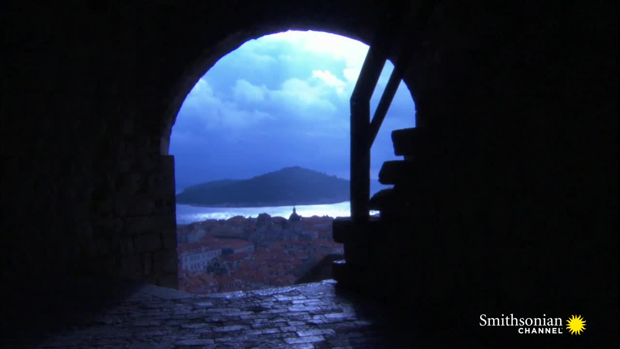 Through the arched opening, the sea and a distant island are visible under a moody, blue sky. Below, a city with red-tiled roofs stretches toward the water.
