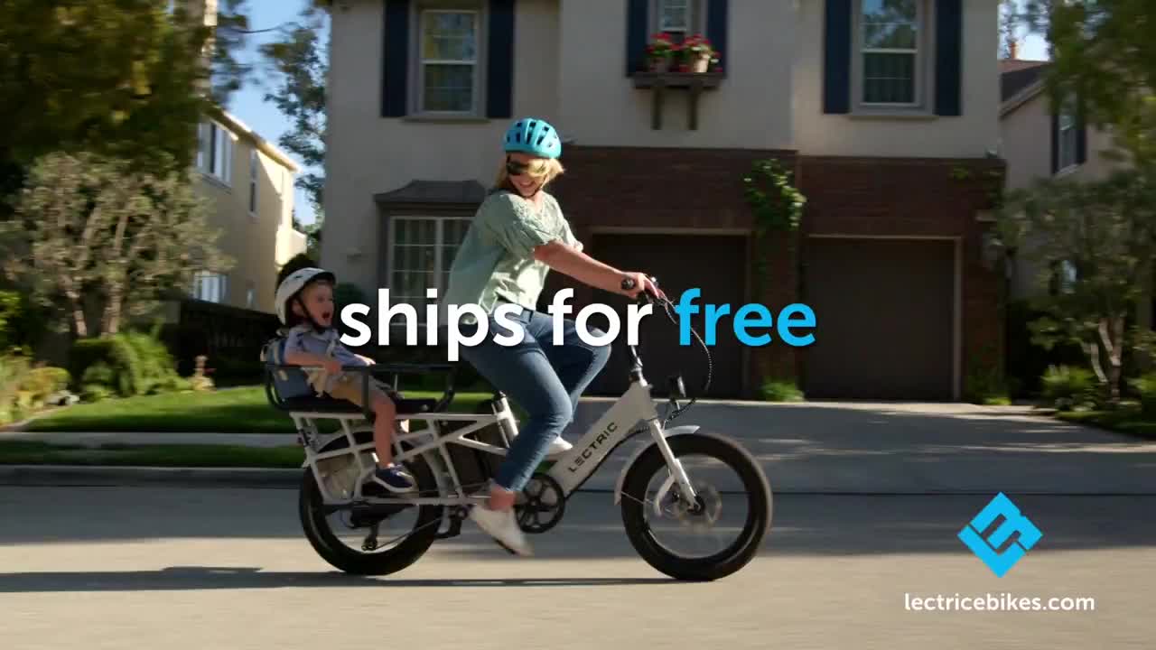 A woman pedals a white electric bike down a tree-lined street, a child secured in a seat behind her. The bike is moving smoothly, with the "ships for free" text superimposed on the scene.
