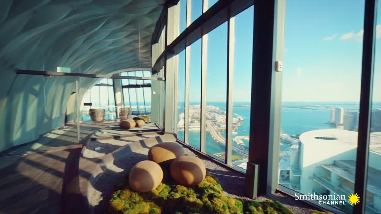 Sunlight streams through the floor-to-ceiling windows, illuminating a modern interior with organic shapes and a view of a cityscape. The Smithsonian Channel logo is visible in the corner.
