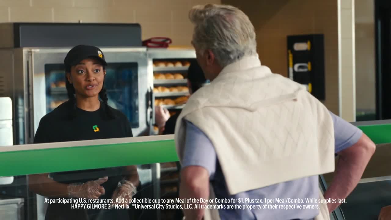 A Subway employee in a black uniform and cap is speaking to a customer. The man, seen from behind, wears a sweater draped over his shoulders.

