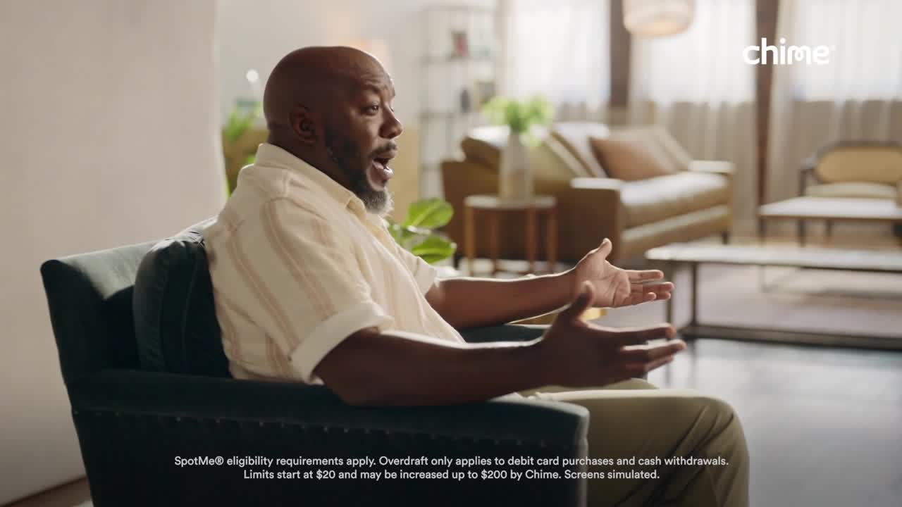 A man sits in a teal armchair, gesturing with both hands as he speaks. His mouth is open, and the sunlight from a nearby window illuminates his face and the neutral-toned living room in the background.
