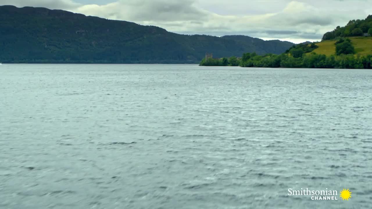 The surface of the lake is rippling slightly, and the overcast sky reflects in the water. Across the lake, a castle sits nestled among trees, as seen on the Smithsonian Channel.
