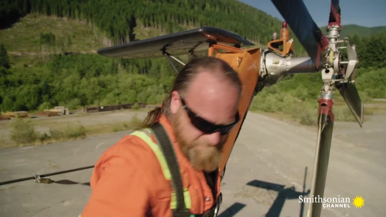 A man in an orange jumpsuit and sunglasses stands next to a helicopter's rotor, the blades still. The scene looks like it's from a show on the Smithsonian Channel, with a backdrop of forested mountains.
