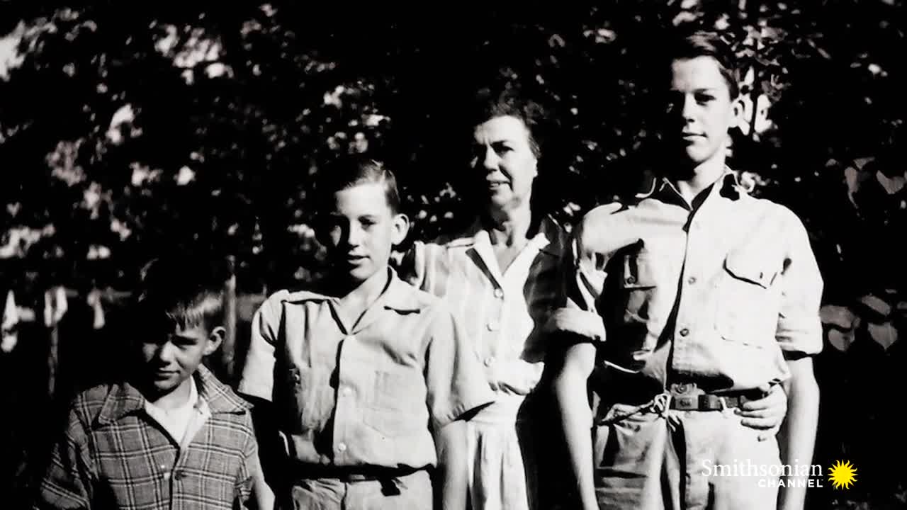 A woman stands with three young men, all posed in front of a dark, leafy background. The Smithsonian Channel logo sits in the lower right corner of the black and white photograph.
