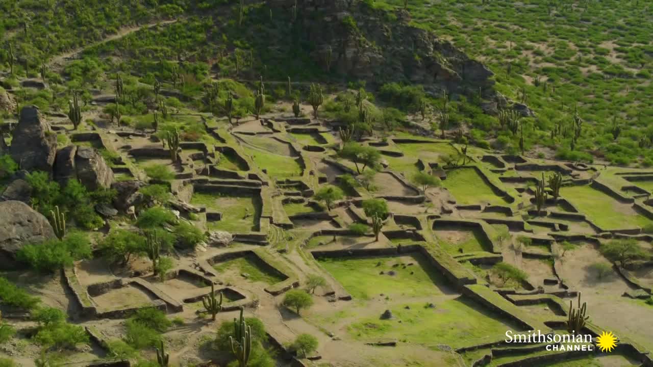 Stone foundations of buildings define a grid across the landscape, with green patches of grass filling the spaces. Tall cacti and small trees dot the area, and the Smithsonian Channel logo is in the corner.
