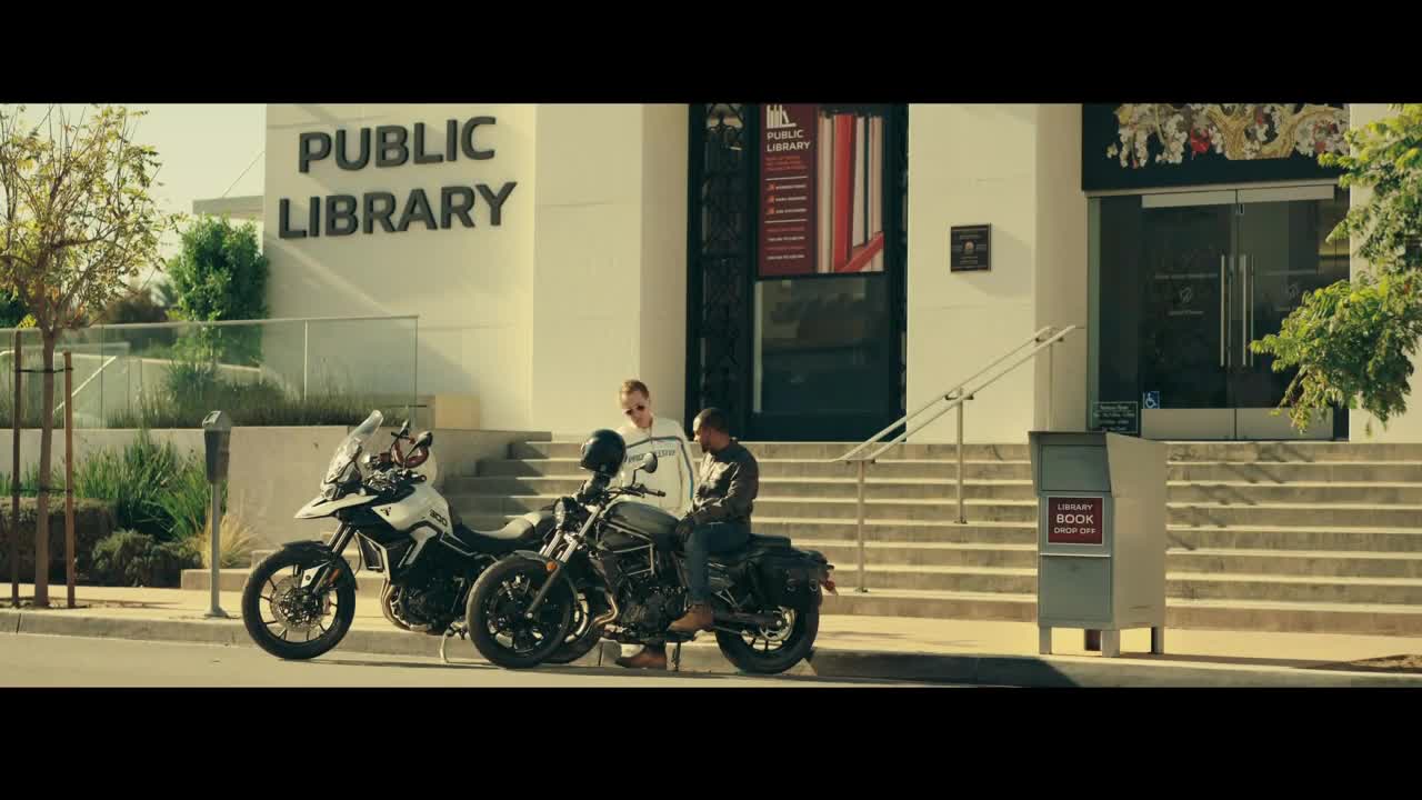 Two motorcycles sit parked in front of a public library. A man sits on one, talking to a woman standing nearby.
