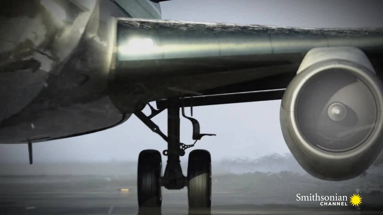 The underbelly of a plane sits low to the ground, its landing gear and engine clearly visible. It's a misty day, and the tarmac reflects the muted light.
