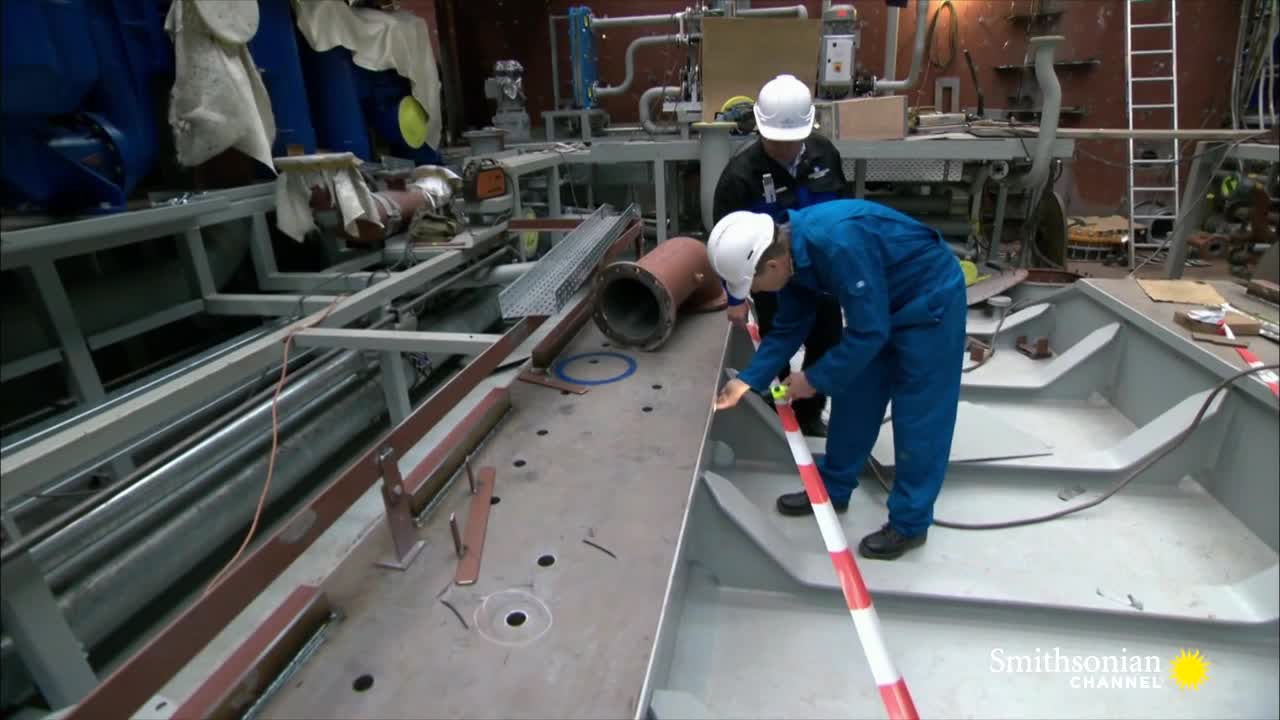 Two workers in blue coveralls and hard hats are measuring inside a metal structure with a red and white striped tape. The scene, from the Smithsonian Channel, takes place in a workshop, with various pipes and metal components scattered around.
