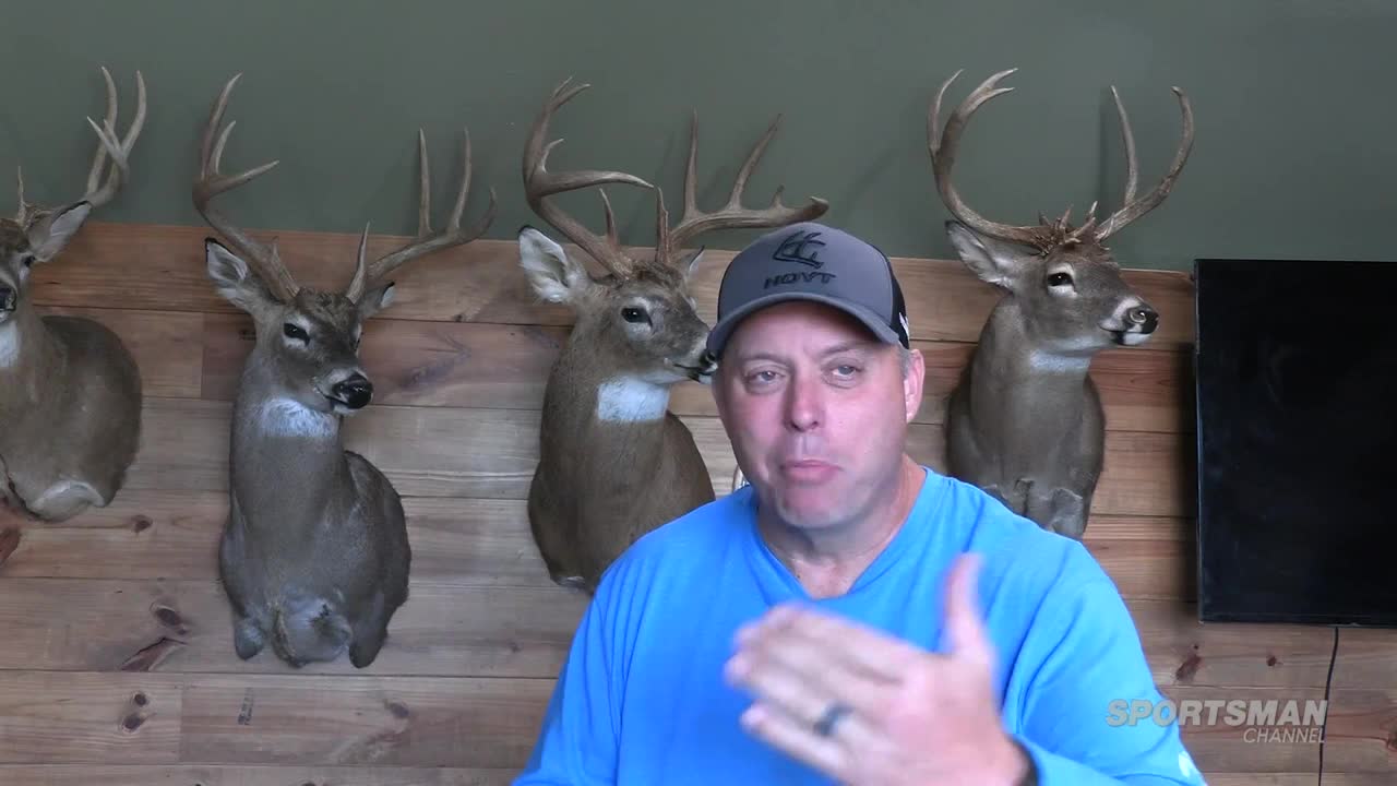 A man in a blue shirt and a cap gestures with his right hand, speaking directly to the camera. Behind him, mounted deer heads hang on the wood-paneled wall, and the Sportsman Channel logo is visible in the lower right corner.
