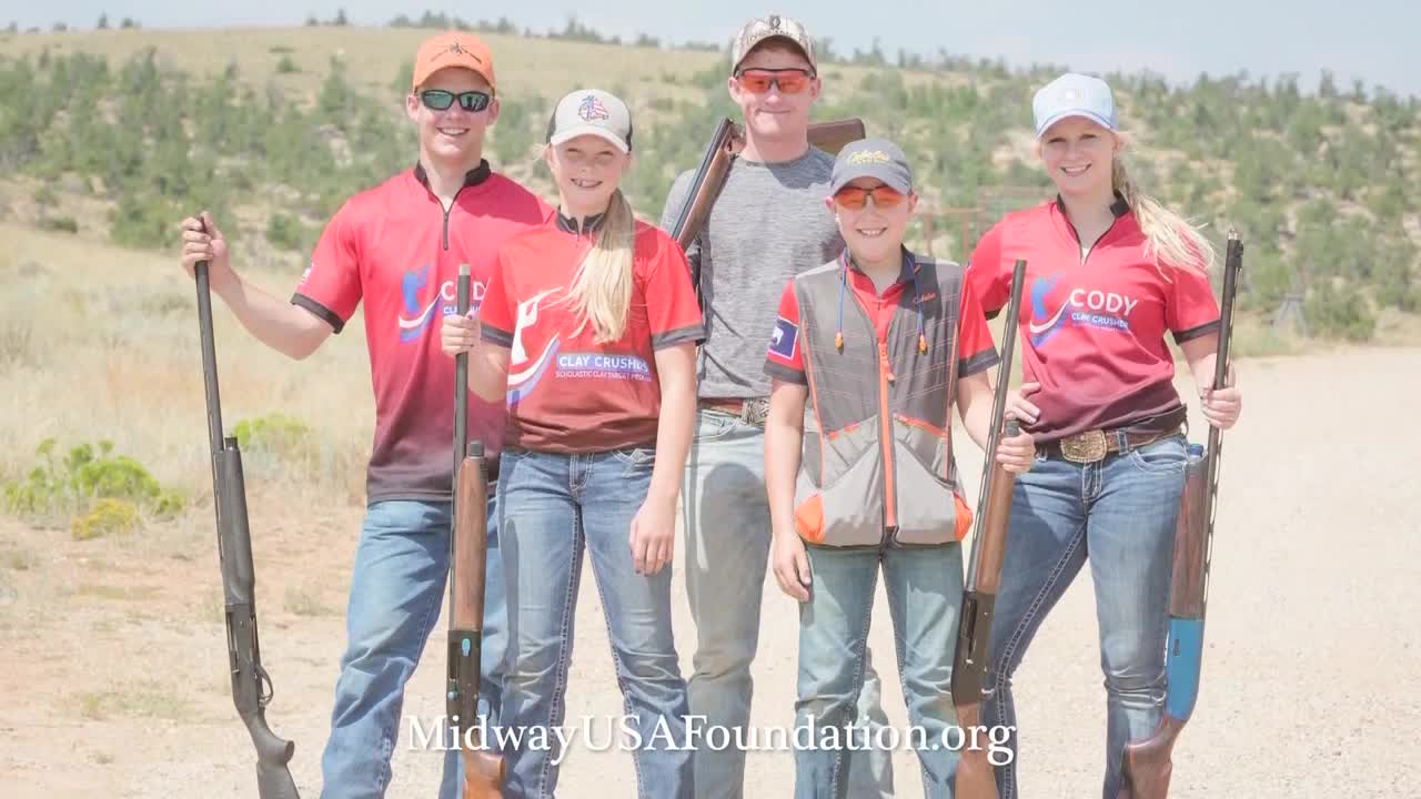 Five people, all wearing shooting apparel, pose together holding shotguns. They're standing on a dirt road, with a backdrop of scrub brush and a bright sky.
