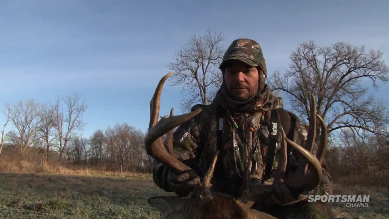 A hunter, clad in camouflage, holds a large set of antlers, the tines curving towards the camera. The Sportsman Channel logo is visible in the corner, and the background shows a field and bare trees under a clear sky.
