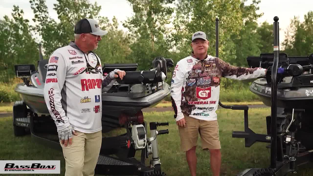 Two men in fishing gear stand next to their bass boats, posing for the camera. The boats are parked on trailers, and the background is a line of green trees.
