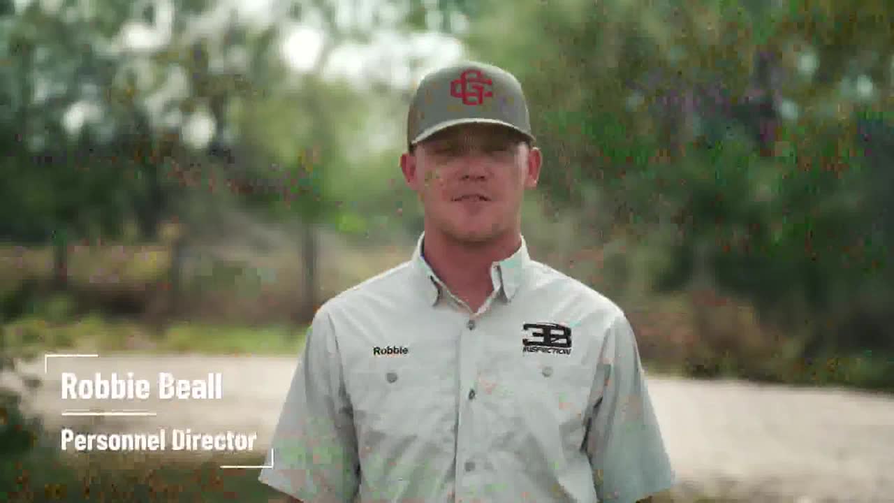Robbie Beall, the Personnel Director, stands in front of a blurred background, likely outside. He's wearing a button-down shirt and a baseball cap, and he's looking directly at the camera.
