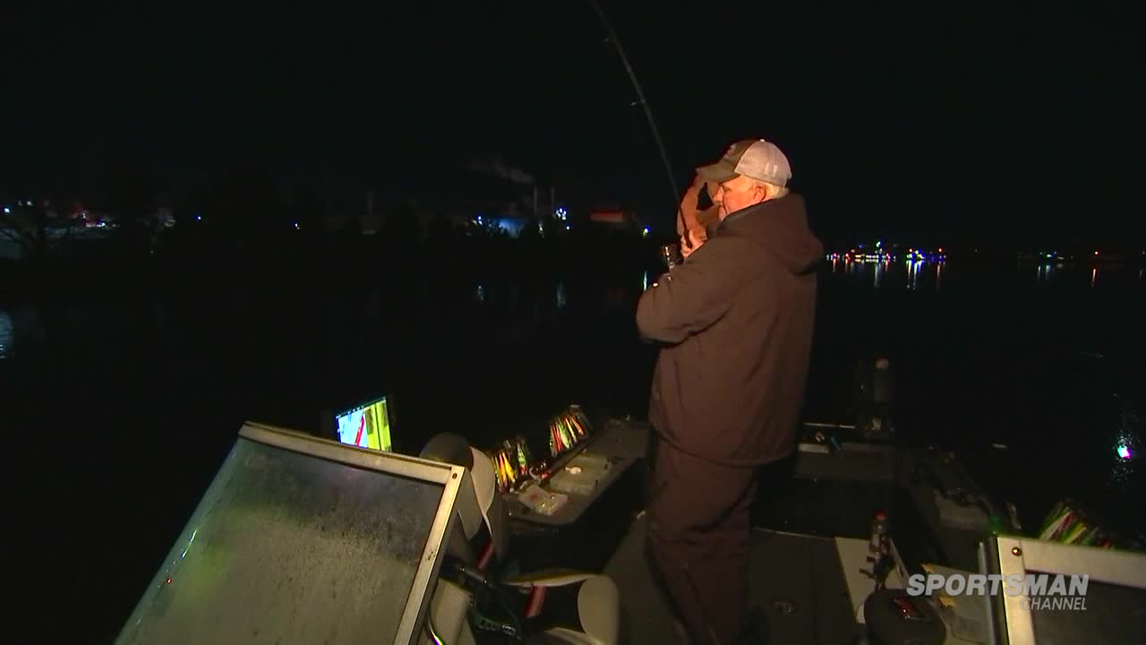 On the water, a man in a cap and jacket is reeling in a fishing rod. The Sportsman Channel logo is visible as he works under the night sky.
