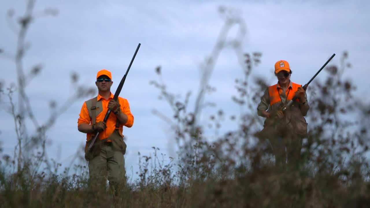 Two hunters, dressed in orange vests and hats, stand in a field, each holding a shotgun. The man on the right carries what appears to be a bird in his hand.

