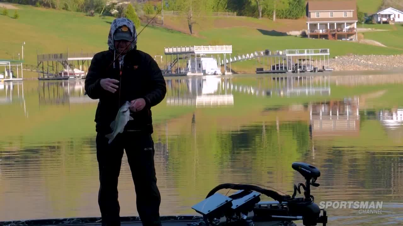 A fisherman, bundled in a jacket and hood, holds up a small fish. The Sportsman Channel logo is visible as he stands on a boat near a calm lake reflecting the shoreline.
