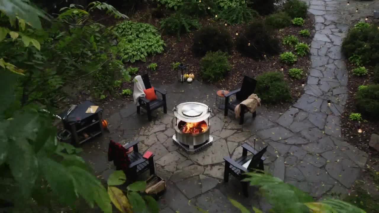Flames dance inside a fire pit surrounded by Adirondack chairs on a stone patio. Overhead, tiny lights twinkle in the surrounding greenery, creating a cozy atmosphere.
