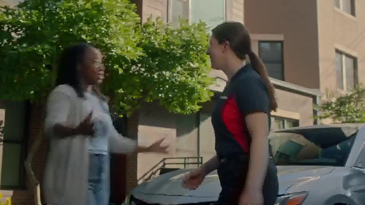 A woman in a gray cardigan gestures animatedly as she speaks to another woman wearing a work uniform. They're standing beside a silver car, with a building and some greenery in the background.
