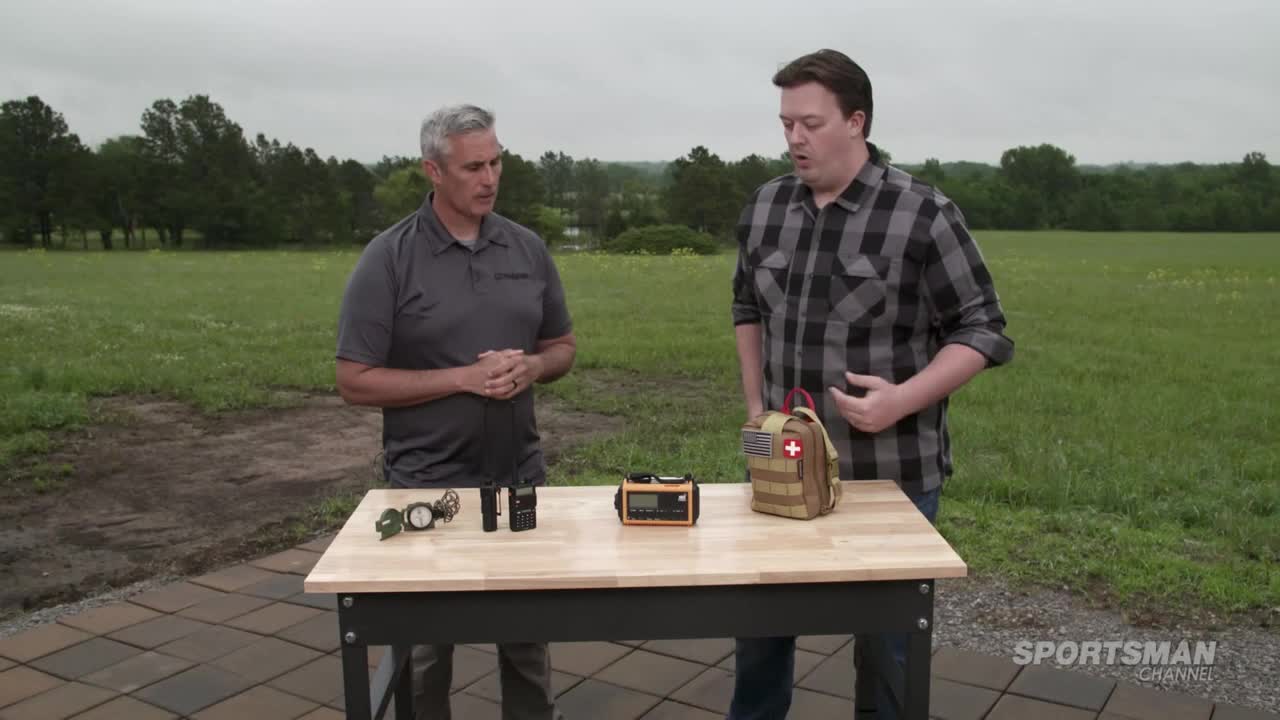 Two men stand behind a table displaying various outdoor gear, including a first-aid kit. The man in the plaid shirt gestures toward the kit while speaking, as the other man listens.

