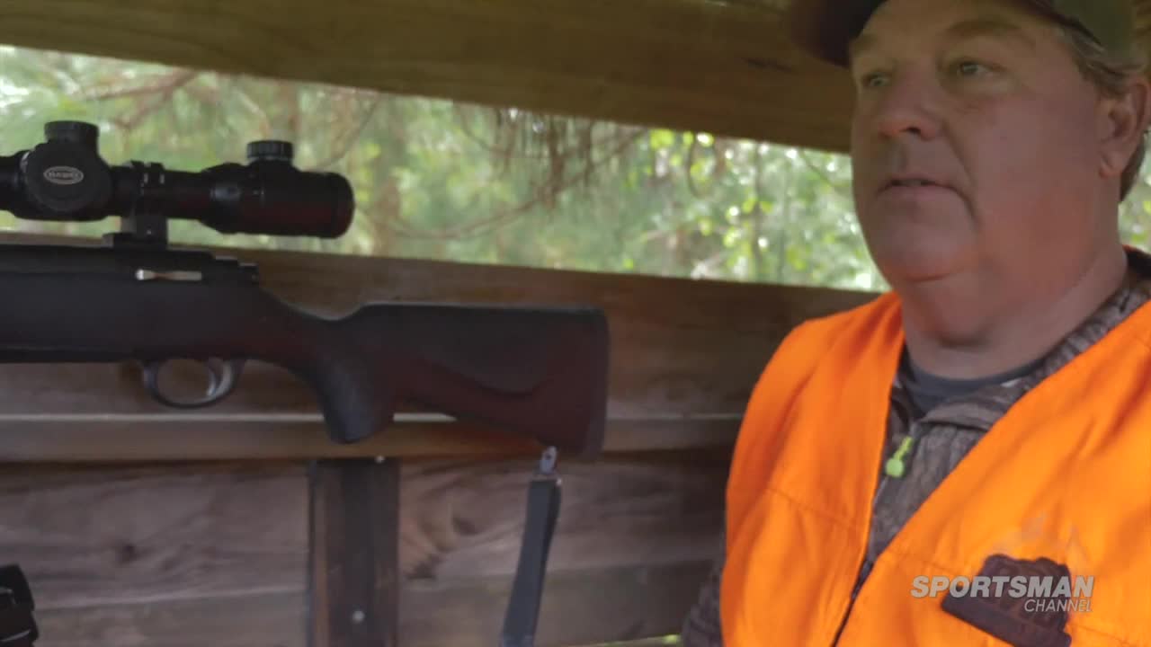 A man in an orange vest sits inside a wooden hunting blind, a rifle with a scope resting beside him. The Sportsman Channel logo is visible on his vest.
