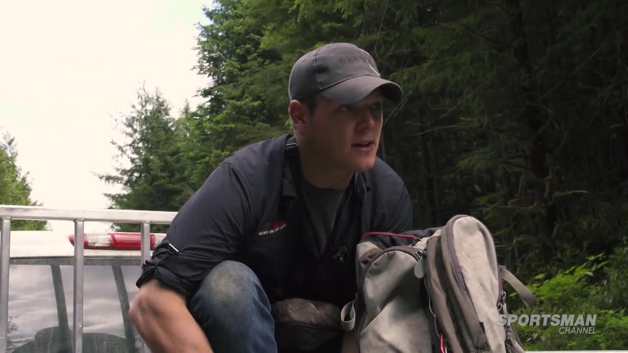 A man in a cap and dark shirt is crouched in the back of a truck, talking. The Sportsman Channel logo is visible, and the scene appears to be filmed in a wooded area.
