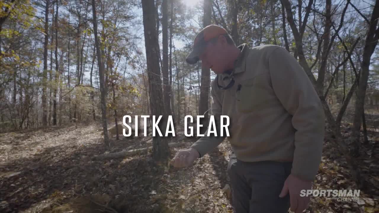 A man in a hunting cap and jacket gestures towards the ground in a sun-dappled forest. The Sportsman Channel logo is visible in the corner.
