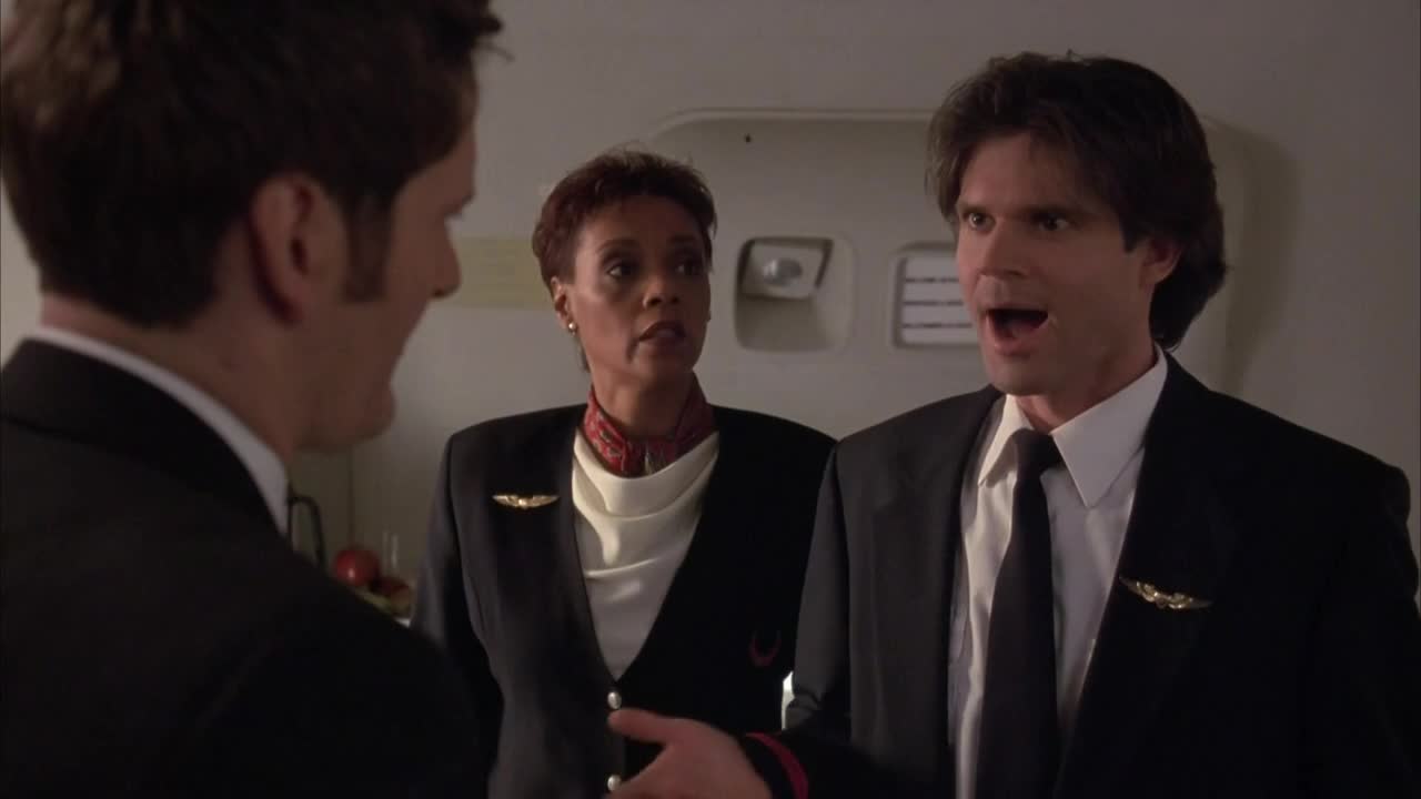 Inside a plane, a man in a suit gestures emphatically while speaking to another man and a flight attendant. The attendant looks on, her expression a mix of concern and professional composure.
