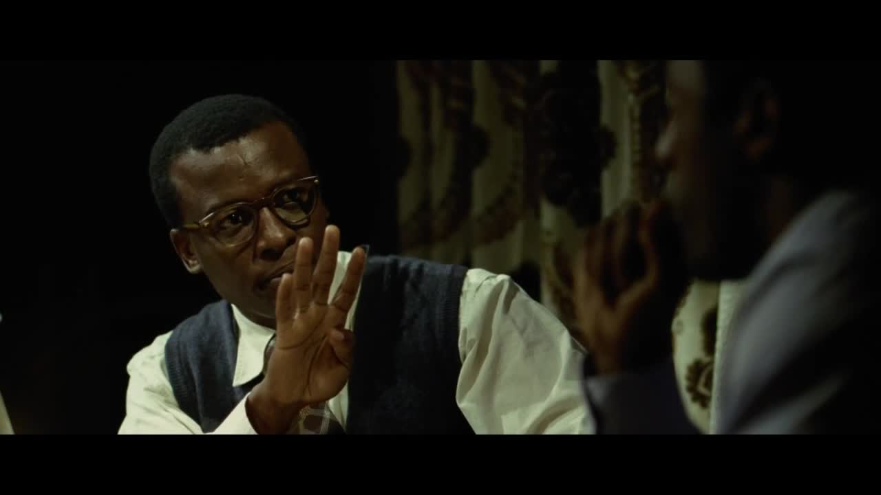 A man in glasses gestures with his right hand, fingers spread, as if making a point. Across from him, another man listens intently, his face partially obscured.
