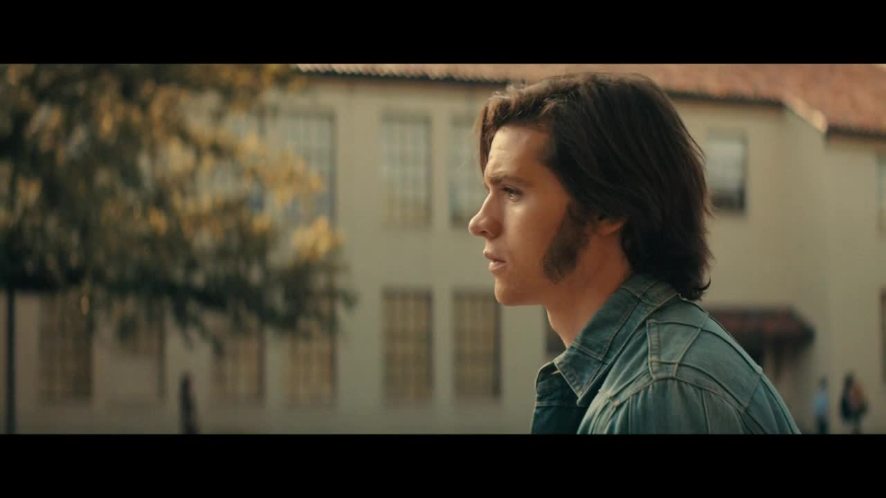 A young man in a denim jacket gazes off-screen, his side profile framed by sideburns. Behind him, a stately building stands in the sunlight, and the scene has a vintage feel, like something from Starz Encore Classic.
