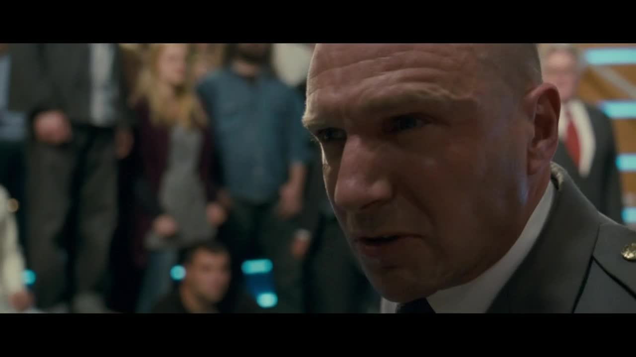 The man in uniform is speaking, his gaze intense. Behind him, a crowd of people stands, their faces mostly obscured.
