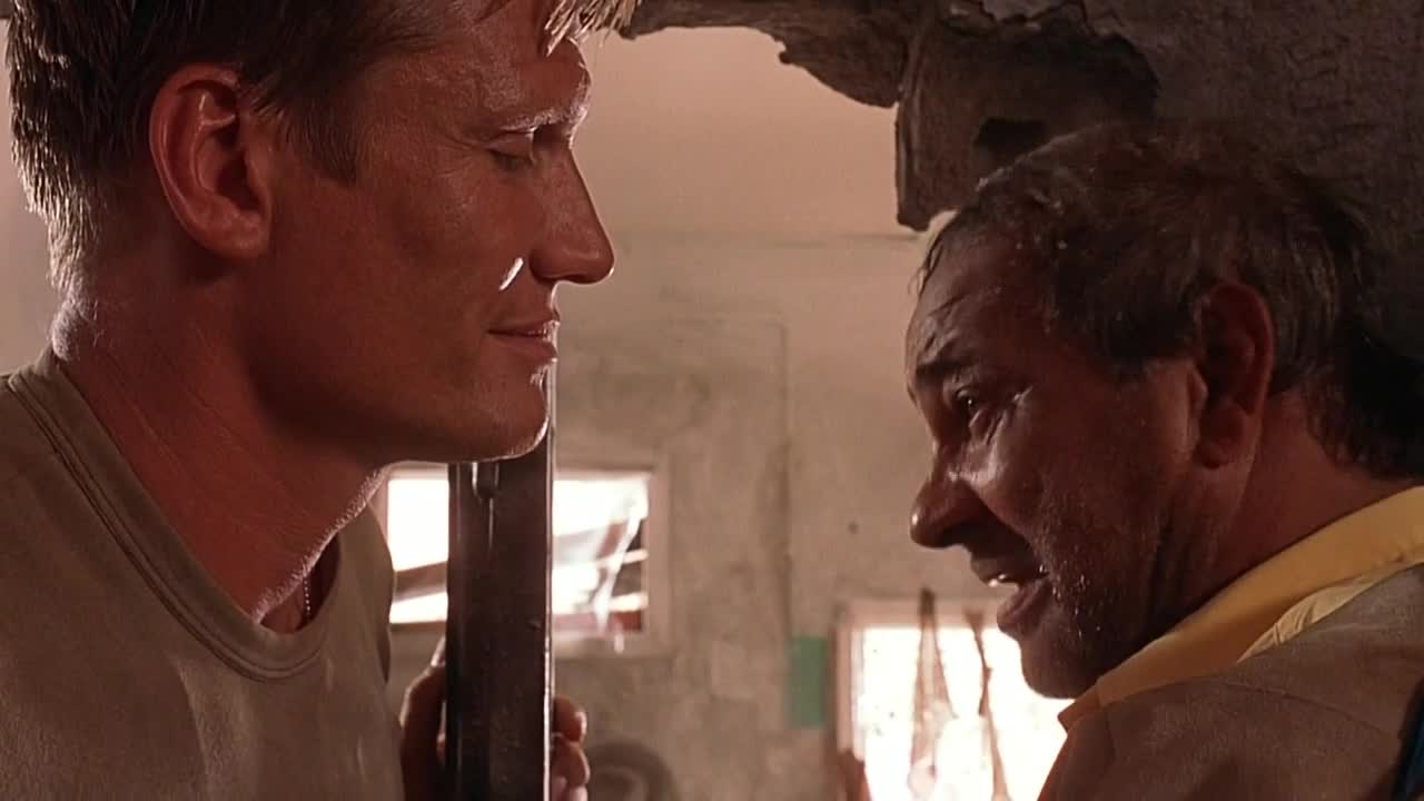Two men, close together, are in a tense conversation. One, with fair hair, looks on, while the other, older, seems to be speaking with a look of concern.
